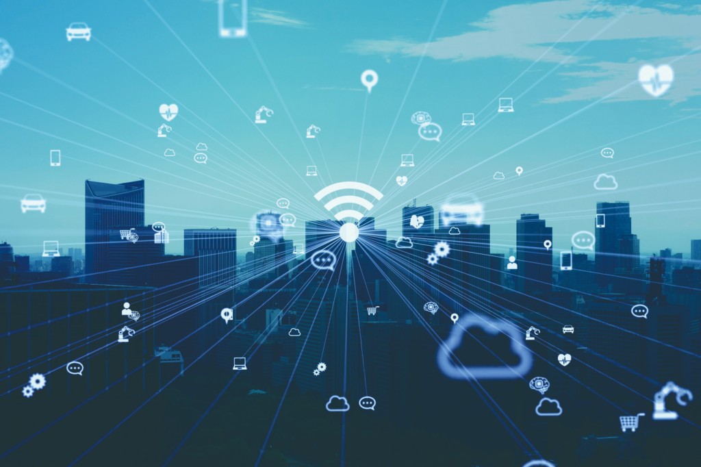 青空の下、ビル群とさまざまなデジタルアイコン（Wi-Fi、携帯電話、医療など）が描かれ、IoTの未来を象徴しています。