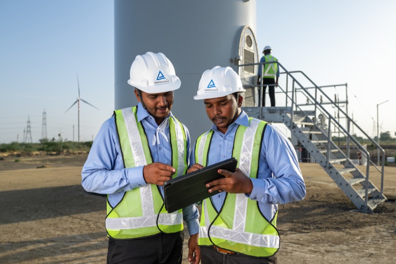 Zwei Ingenieure in Sicherheitswesten und Schutzhelmen mit einem Tablet vor einer Windkraftanlage