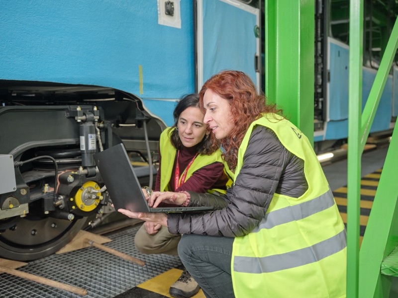 Two engineers in safety vests reviewing data on a laptop next to rail vehicle components in a workshop