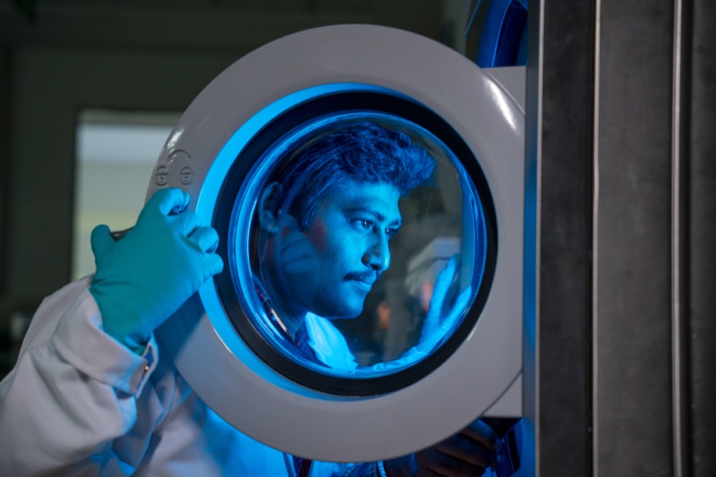 Laboratory technician looking through the round window of a testing machine illuminated in blue light