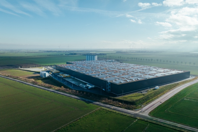 Aerial view of a large industrial building with solar panels on the roof surrounded by green fields